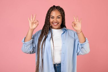 Neşeli Siyah Artı Boyutlu Kadın Hareketi. Pembe arka planda stüdyoda dikilirken iki eli kameraya gülümseyen 