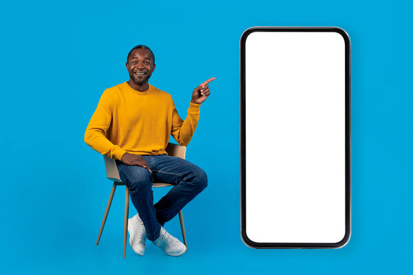 Relaxed handsome middle aged black man in casual sitting on chair over blue studio background, pointing at huge smartphone with white blank screen, showing entertaining mobile app, mockup