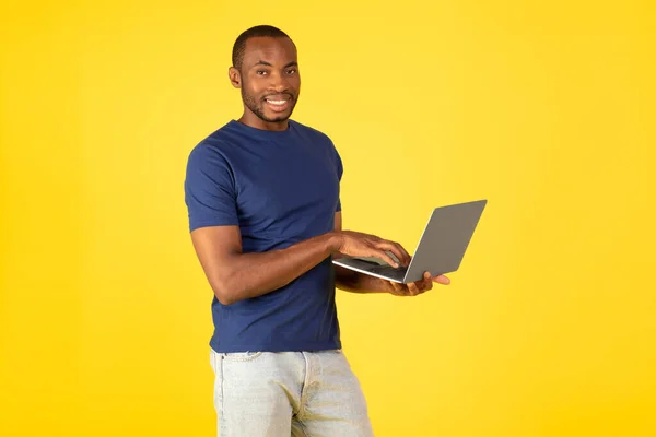 黄色のスタジオの背景に立ってコンピュータを保持ポーズカメラにノートパソコンの笑顔を使用してハッピーブラックマン オンラインとウェブサーフィンで働く男 インターネット技術 — ストック写真