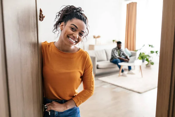 Welcome To Our Home. Cheerful Black Wife Meeting You In Opened Door Smiling To Camera Standing While Husband Sitting In New Apartment. Real Estate Purchase Concept. Selective Focus