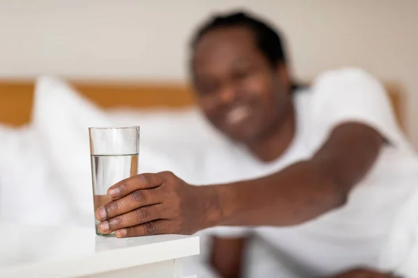 Black Smiling Man Taking Glass With Water From Bedside Table In The Morning, Happy Young African American Guy Reaching Hand To Refreshing Mineral Drink While Relaxing In Bed, Selective Focus
