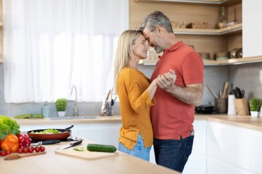 Romantik orta yaşlı çift, günlük giysiler içinde mutfakta yemek pişirirken, dans ederken, birbirlerine bakarken, gülümserken, mutlu bir koca, güzel karısına sarılırken, mekanı kopyalarken.
