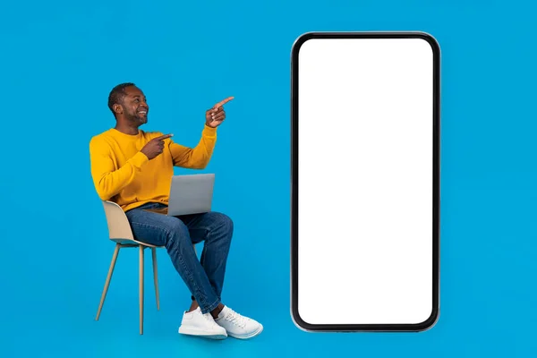 身穿休闲装的积极的非洲中年男人坐在椅子上 膝上型计算机 指着巨大的手机 屏幕是白的 背景是蓝的 是个模仿者 — 图库照片