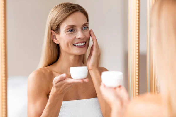 顔の皮膚治療の概念 バスルームで鏡の前にタオルで包まれた顔に保湿クリームを適用する中年女性 — ストック写真