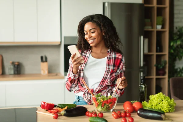 예쁘고 아프리카 미국인 아가씨가 샐러드를 준비하고 유기농 야채와 내부의 스마트폰에서 — 스톡 사진