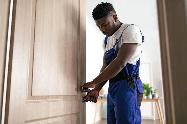 Doors Installation Service. African American Professional Repairman Fixing Door Lock For House Safety Standing Wearing Blue Coverall Uniform At Home. Renovation Concept