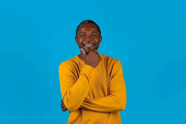 快乐而又英俊的非洲男子 穿着黄色毛衣 独自站在蓝色工作室的背景上 触摸着下巴 高兴地对着相机微笑 复制空间 — 图库照片
