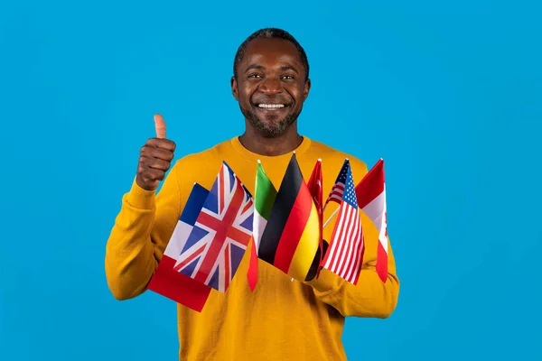 Happy middle aged african american man in yellow posing on blue studio background, holding various flags of different countries and showing thumb up, copy space. Foreign language school, immigration