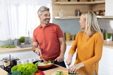Neşeli orta yaşlı eşler birlikte yemek pişiriyor ve sohbet ediyorlar, güzel sarışın kadın sebze kesiyor ve yakışıklı kocasıyla sohbet ediyor yemek yapıyor, mutfak içi, fotokopi odası.