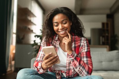 Akıllı telefonda yazan, sosyal medyada tek başına sohbet eden, oturma odasındaki koltukta oturan, gülümseyen Afro-Amerikan bir bayan. Blog, iş ve mesaj evde, yeni uygulama, reklam ve vadesi covid-19