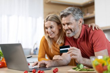 Sevgi dolu bir çift yemek pişirirken internetten sipariş veriyor, mutfakta duruyor, modern laptop ve kredi kartı kullanıyor, internetten yemek alıyor, fotokopi çekiyor. Kolay alışveriş kavramı