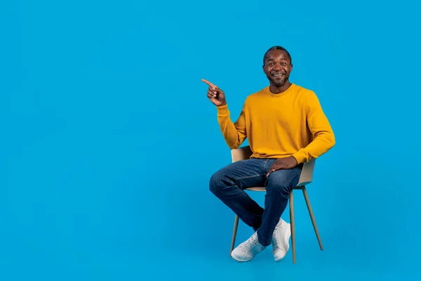 快乐英俊的中年黑人男子坐在蓝色工作室的椅子上 指着广告和微笑的复制空间 显示出很高的出价 全长镜头 — 图库照片