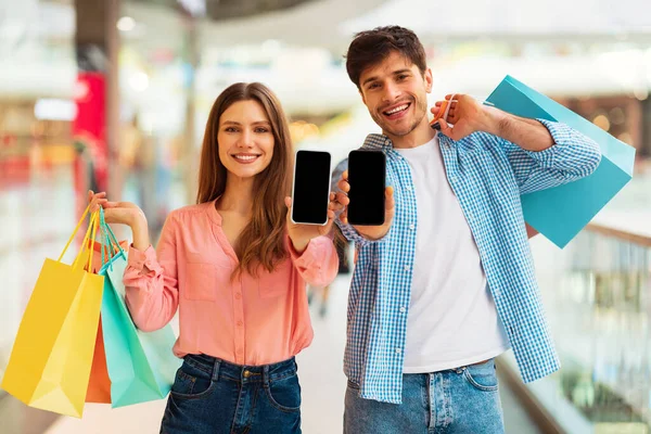 Mobile Shopping App. Happy Buyers Couple Showing Cellphones Advertising App Smiling To Camera Holding Shopper Bags Posing In Modern Mall Indoor. Ecommerce And Technology Concept. Mockup