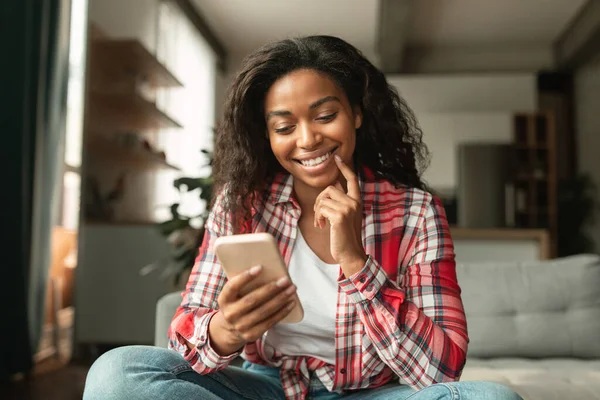 Smiling pretty young african american lady typing on smartphone, chatting in social media alone, sit on sofa in living room interior. Blog, work and message at home, new app, ad and offer due covid-19