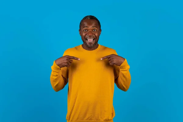 Are you talking about me. Excited handsome middle aged african american man, looking surprised pointing himself, making sure he heard right, being chosen, posing over blue background