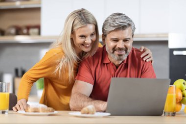 Mutfakta kahvaltı yaparken dizüstü bilgisayar kullanan mutlu eşler taze meyve suyu içen, meyve ve pasta yiyen, internetten alışveriş yapan, yeni mobilya seçen, fotokopi çeken kocalar.