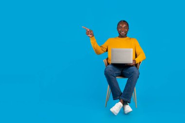 Pozitif orta yaşlı Afro-Amerikalı sıradan bir adam mavi stüdyo arka planında koltukta oturuyor, online çalışıyor, modern dizüstü bilgisayar kullanıyor, fotokopi alanını işaret ediyor, tam boy.