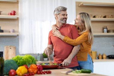 Gülümseyen orta yaşlı koca karısı için lezzetli sağlıklı yemekler hazırlıyor, mutlu sarışın kadın eşine sarılıyor yemek pişiriyor, mutfak içi, yan manzara, fotokopi odası.