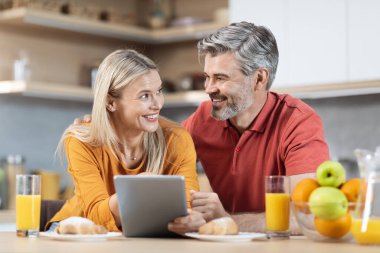Mutlu orta yaşlı çift evde sağlıklı bir kahvaltı yapıyor, masada oturuyor, taze portakal suyu içiyor, meyve yiyor, dijital tablet kullanıyor ve gülümsüyor.
