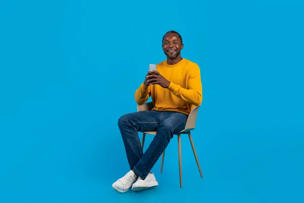 宽松的中年黑人男子 身穿休闲装 坐在蓝色工作室的椅子上 使用手机 与朋友聊天 复制空间 全身而退 — 图库照片