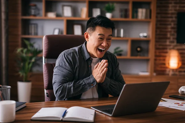 Happy adult japanese man look at laptop, gesturing and greeting, have video call with client in home office interior. Businessman communicating remotely, say hi. Social distance and modern business