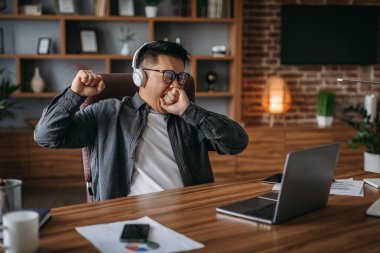 Tired boring adult korean man manager in glasses and headphones yawns and rests from work in home office interior. Development, business and social distancing, take break, deadline and client support