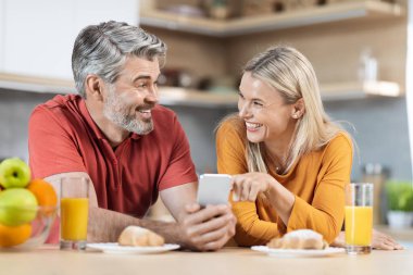 Neşeli olgun eşler hafta sonu kahvaltısını planlarken mutlu orta yaşlı karı koca mutfak masasında oturup atıştırıyor ve akıllı telefon kullanıyor, otel rezervasyonu yapıyor.