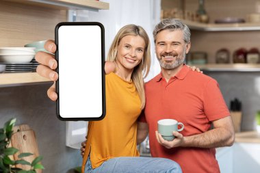 Evde birlikte kahve içen, beyaz ekranlı ve gülümseyen, mobil uygulama reklamı için model, mutfak iç mekanı olan akıllı telefon gösteren orta yaşlı, pozitif bir çift.