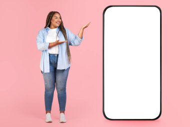 Siyah kilolu kadın, Pembe Arkaplanda Stüdyoda Mobil Uygulama Pozisyonunu Boş Ekran Tavsiyesi Veren Büyük Akıllı Telefon Gösteriyor. Mockup, Tam Uzunluk Çekimi