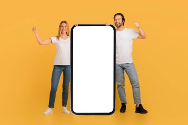Beyaz tişörtlü, tatmin olmuş beyaz tenli genç çift, sarı arka planda izole edilmiş uygulamayı onaylayan, boş ekranlı, kocaman bir akıllı telefonun yanında baş parmak gösteriyorlar. Aygıt sunumu ve tavsiyesi, reklam ve teklif