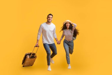 Sarı arka planda bavulla atlayan neşeli genç Arap çiftin portresi, saman şapkalı mutlu Orta Doğulu adam ve kadın gülüyor ve kameraya gülümsüyor, seyahat ve tatilin tadını çıkarıyor.