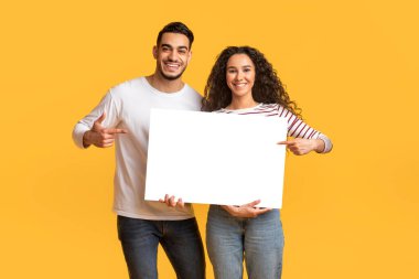 Reklam için yer. Ellerinde boş beyaz plaketi tutan ve gülümseyen Orta Doğulu genç erkek ve kadın reklam için kopya alanı gösteren mutlu bir Arap çifti.