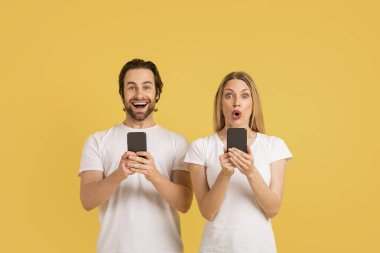 Beyaz tişörtlü, ağzı açık, akıllı telefonlarda sohbet eden, sarı arka planda, stüdyoda izole edilmiş neşeli beyaz bir çift. Harika haberler, sosyal medya, uygulama, reklam ve zafer duyguları