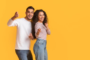 Olumlu genç Arap erkek ve kadın kamerayı işaret ediyor, neşeli orta doğulu çift, sarı arka planda tek başına dikilip, uzayı kopyalayarak birini işaret ediyor.