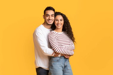 Kameraya sarılan ve gülümseyen genç Arap erkek ve kadının portresi, sarı arka planda birlikte poz verirken romantik Orta Doğu çifti,