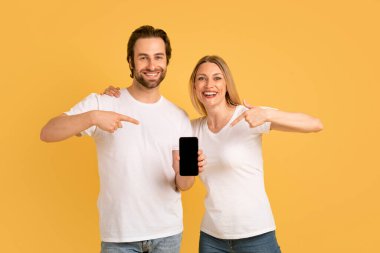 Beyaz tişörtlü mutlu beyaz beyaz tenli çift akıllı telefonu boş ekranla gösterip sarı arka planda ve stüdyoda izole edilmiş uygulama ve web sitesini tavsiye ediyor. Reklam ve teklif, online zafer