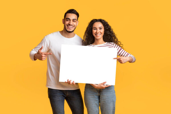 Place For Ad. Happy Arab Couple Holding And Pointing At Blank White Placard In Their Hands, Smiling Young Middle Eastern Man And Woman Demonstrating Copy Space For Advertisement, Mockup