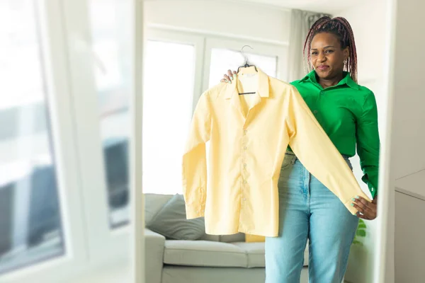 African american lady choosing clothes to wear, looking herself in mirror in living room at home, free space. Black woman thinking what to wear. Lifestyle women relax at home concept.