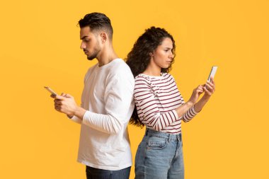 Gadget Bağımlılık Konsepti. Sırt sırta duran ve akıllı telefonlar kullanan genç bir Arap çift Orta Doğulu erkek ve kadın cep telefonlarıyla zaman geçirirken birbirlerini görmezden geliyorlar.