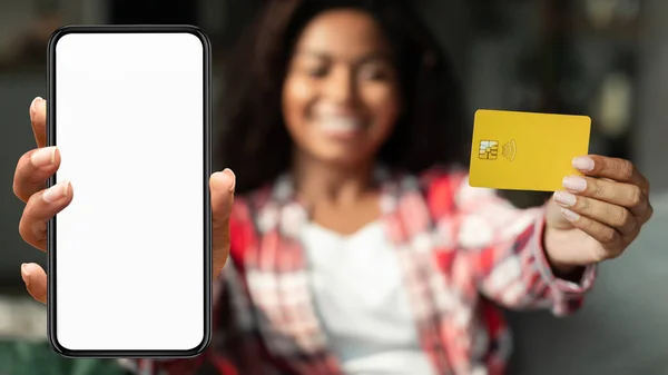 Happy millennial black female show credit card, smartphone with blank screen in living room interior. Woman enjoy online shopping at home, purchase order remotely and social distancing, app banking