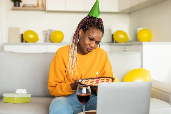 Online party. Sad single black woman in party hat celebrating b-day online, holding pie and looking at laptop screen. Unhappy lady video calling, sitting on sofa at home