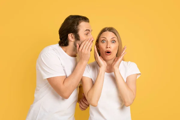 Cheerful young caucasian guy whispering into ear of shocked woman in white t-shirt, isolated on yellow background, studio. Human emotions, gossip, news, great ad and offer, love and relationship