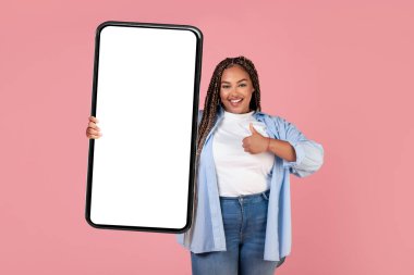 Beyaz ekranlı Büyük Akıllı Telefon 'u gösteren ve parmaklarını kaldırıp pembe arka planda poz veren mutlu Afrikalı Amerikalı kadın. Mobil uygulama kavramını sevdim. Model