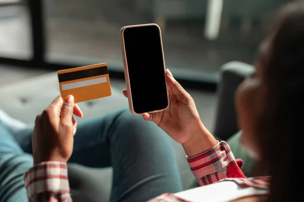 Smiling young african american lady show credit card and smartphone with blank screen in living room interior. Online shopping at home alone, purchase order remotely and social distancing, app banking