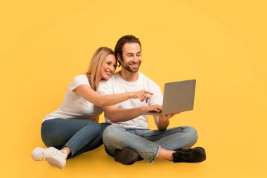 Beyaz tişörtlü, gülümseyen, beyaz tişörtlü adam bilgisayarda, yerde, sarı arka planda, stüdyoda izole bir şekilde oturuyor. Harika reklam ve teklif, online satış, aşk ve ilişki, video görüşmesi.