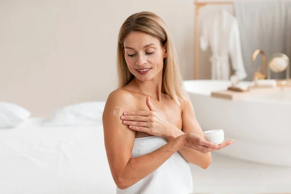 Body treatments. Happy middle aged woman applying moisturising cream on shoulder, sitting wrapped in towel and making beauty care routine at home, copy space