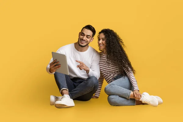 快乐的阿拉伯年轻夫妇 带着数字标签在黄色工作室的背景上在地板上放松 用现代设备在网上购物或浏览互联网微笑着中东男人和女人 — 图库照片