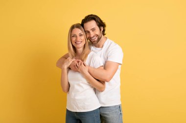 Beyaz tişörtlü kadını kucaklayan, birlikte romantizmin tadını çıkaran, sarı arka planda, stüdyoda izole edilmiş yakışıklı beyaz adam. İyi reklam ve teklif, aşk, ilişki ve romantizm boş vakitlerde