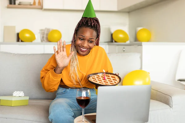 Remote birthday party. Happy african american lady holding pie and video calling friends on her birthday, celebrating online, waving hand to laptop and smiling