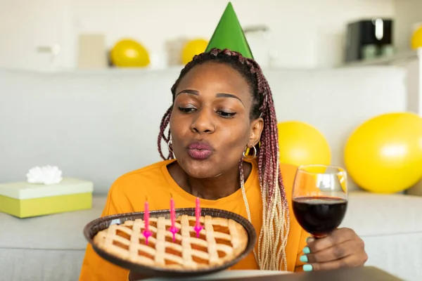 African american woman blowing candles on b-day pie, having online birthday party, wearing festive hat while celebrating at home via video call. Modern distance holiday celebration concept